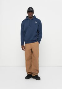 Marine hoodie met bijpassende zak aan de voorkant en een trekkoord, gedragen met beige cargo shorts en zwarte sneakers, gecombineerd met een zwarte pet met een logo.