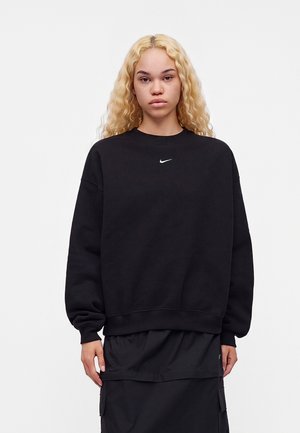 Młoda osoba z długimi blond kręconymi włosami, ubrana w prostą czarną bluzę Nike i czarną spódnicę, stoi na białym tle.