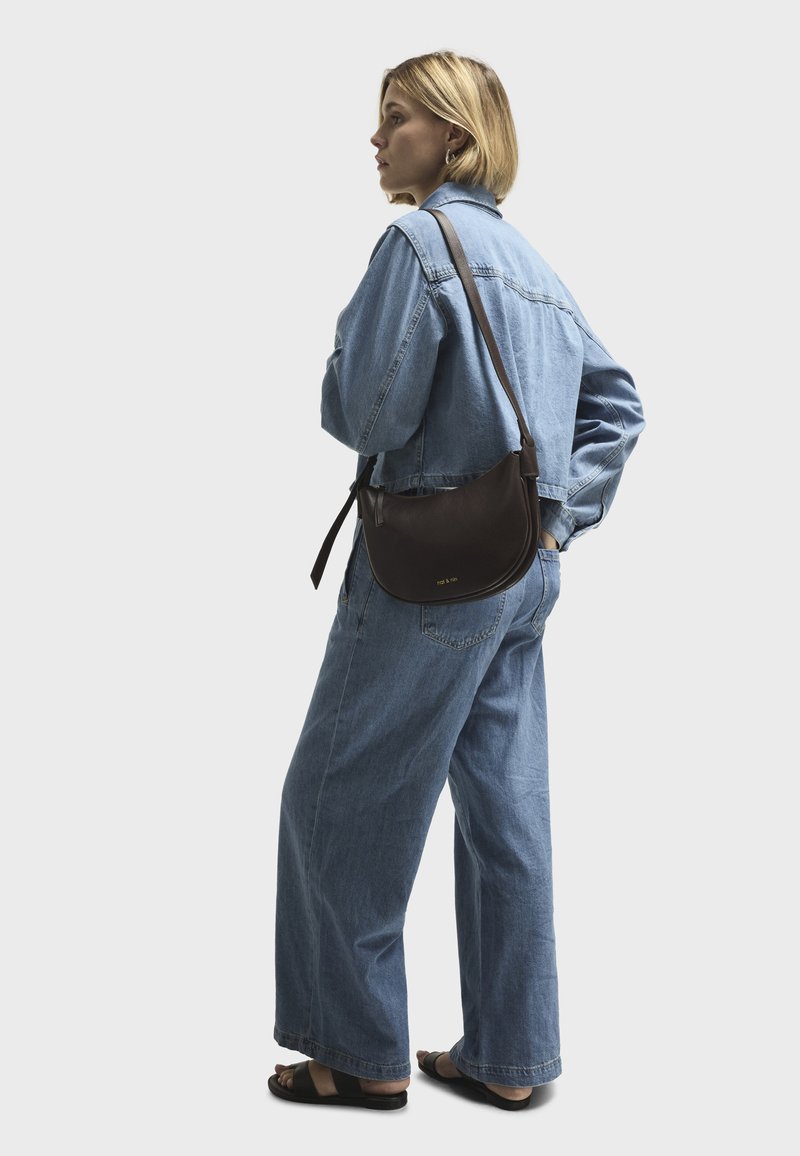 Vrouw in lichtblauwe spijkerjasje en wijd uitlopende jeans, met een bruine leren schoudertas, staand met één hand in haar zak.