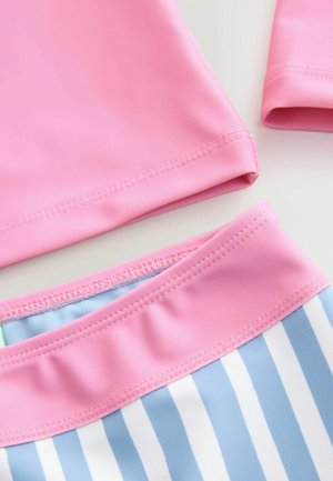 Różowa bluzka z długim rękawem o gładkiej fakturze, zestawiona z niebiesko-białym paskiem wzdłuż talii, z falbaniastym wykończeniem na pasku.