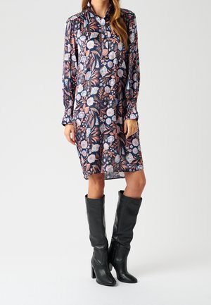 Femme portant une robe à manches longues fleurie avec des fleurs roses et bleues, associée à des bottes en cuir noires montantes à talons.