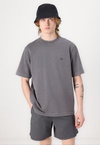 Šedé oversized tričko s krátkým rukávem a malým logem, doplněné o šedé šortky. Na hlavě má černý klobouk s přezkou.