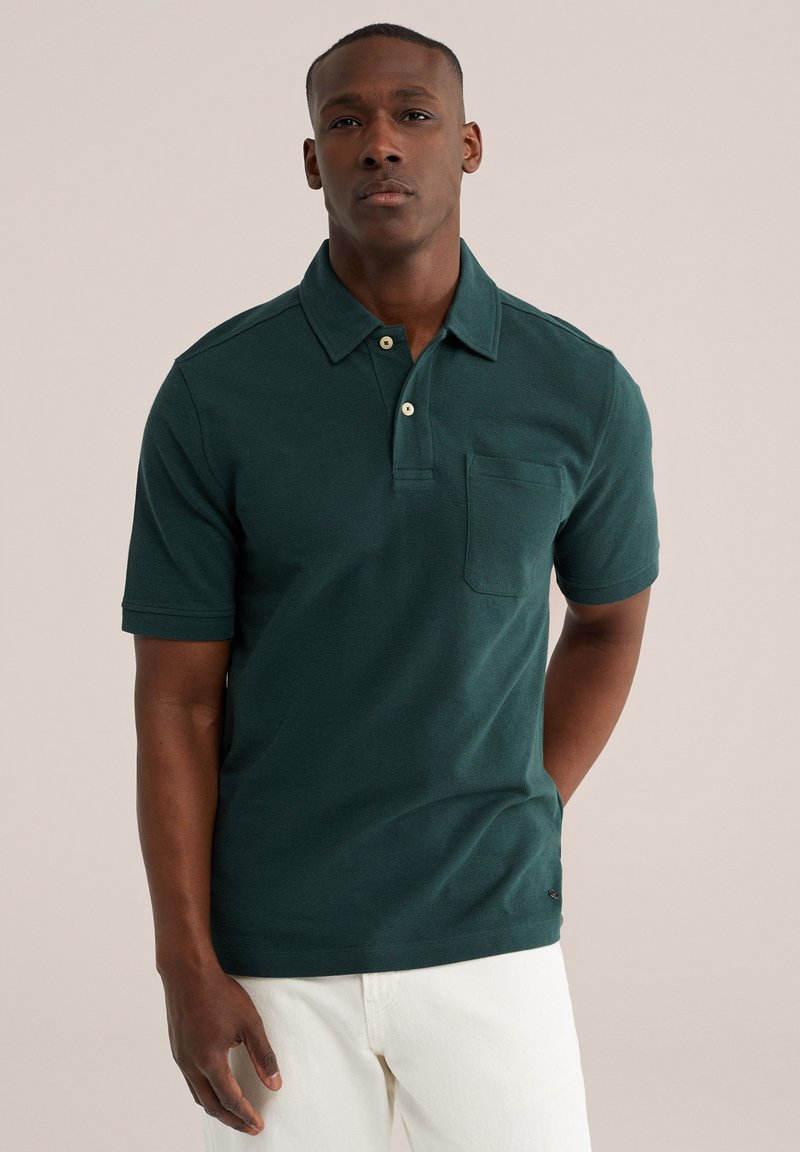 Homme portant un polo vert foncé à manches courtes avec une poche avant et un pantalon blanc, debout devant un fond beige uni.