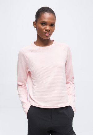 Femme aux cheveux courts portant une chemise rose clair à manches longues et un pantalon noir, debout les mains dans les poches devant un fond uni.