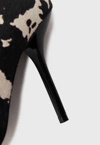 Primer plano de un tacón de aguja negro con tela de estampado de vaca en negro y beige sobre fondo blanco.
