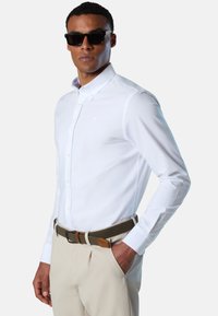 Camicia bianca con bottoni e colletto, con motivo a quadretti all'interno. Abbinata a pantaloni beige e una cintura verde intrecciata. Occhiali da sole neri.