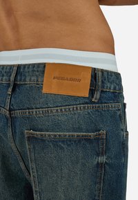 Κοντινό πλάνο της κάτω πλάτης ενός ατόμου που φοράει μπλε τζιν από τζιν με καφέ δερμάτινη ετικέτα «PEGADOR» και άσπρο εσώρουχο με λευκή μέση.