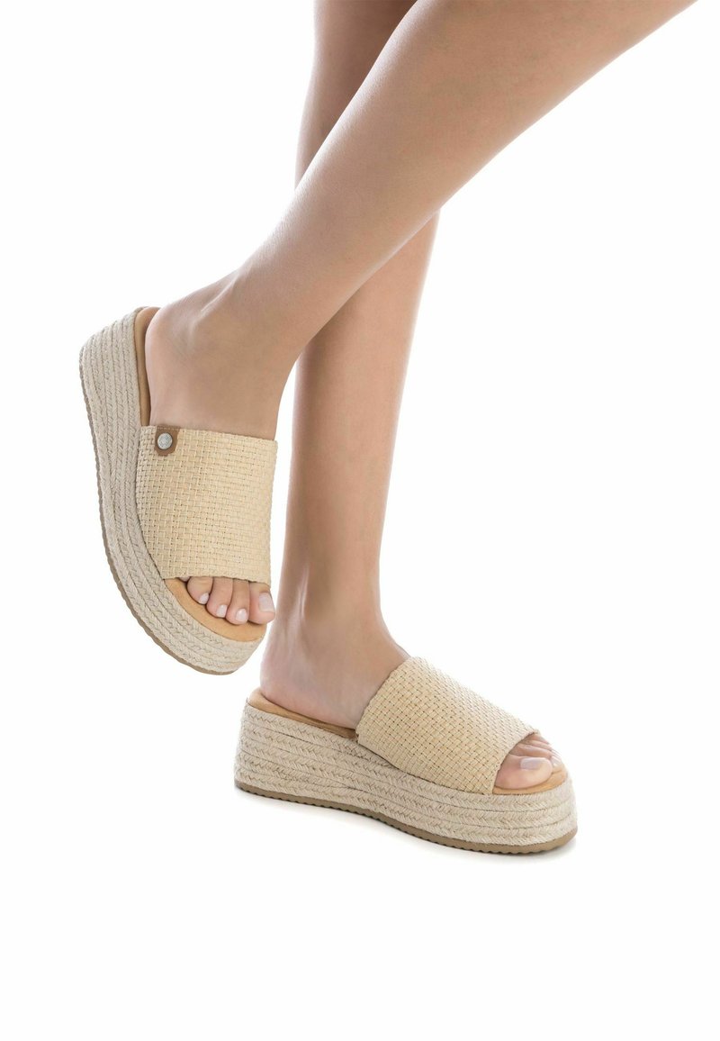 Sandalias deslizantes tejidas en beige con punta abierta, parte superior texturizada y suela gruesa de capas, con un acento metálico circular en el lateral.