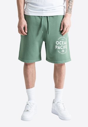 Mand iført grønne shorts med snøre og hvid tekst ”Ocean Pacific”, hvide sokker og hvide sneakers, der viser en tatoveret underarm.