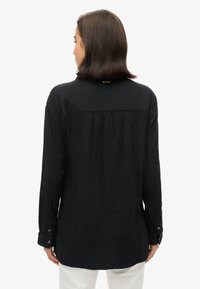 Zwarte shirt met lange mouwen van gestructureerde stof, met knopenmanchetten en een rugpasdetail. Heeft een rechte snit en een afgeronde zoom.