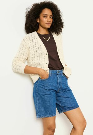 Woman with curly hair wearing a cream knit cardigan, dark brown top, blue denim shorts, and gold necklace, standing with hands in pockets.
