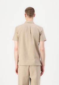 Camisa de botones de manga corta en color beige con tejido texturizado, dobladillo recto y detalle de pliegue en la parte trasera. Llevada con pantalones beige a juego.
