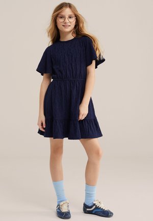 Deklica z dolgimi blond lasmi, ki nosi okrogle očala, mornarsko modro obleko s kratkimi rokavi, svetlo modre nogavice in modre superge, stoji.