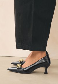 Crne cipele na visoku petu od lakirane kože sa šiljastim vrhovima, ukrašene zlatnim lancem preko prednjeg dijela, prikazane uz crne skraćene hlače.