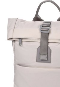 Mochila crema acolchada con tapa plegable, correa gris y herrajes, que cuenta con una cremallera y un bolsillo frontal para almacenamiento extra.