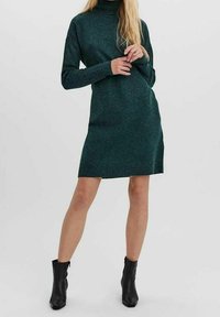 Robe tricotée turquoise avec un col haut, des manches longues et une fente latérale, associée à des bottines noires. Texture lisse et forme ajustée.