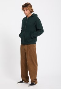 Homme aux cheveux mi-longs portant un sweat à capuche zippé vert foncé, un pantalon en velours côtelé marron et des chaussures noires sans lacets, debout les mains dans les poches.