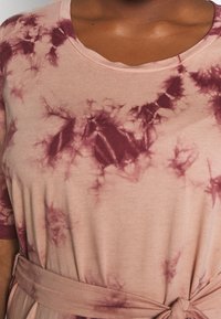 Vaaleanpunainen taitaustoppi, jossa on pyöreä pääntie, lyhyet hihat ja solmittu vyötäröyksityiskohta tumman viininpunaisilla yksityiskohdilla sekä pehmeä rakenne.