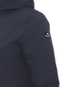 Giacca blu navy con una consistenza liscia e opaca; presenta un logo sulla spalla sinistra e una zip frontale sul lato destro.
