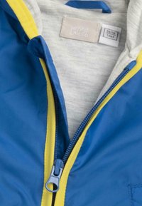 Veste pour bébé bleu et jaune avec une fermeture éclair partiellement ouverte révélant une doublure intérieure grise et une étiquette de taille indiquant 56/1M pour 1 mois.