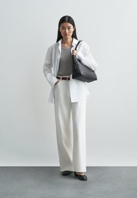 Witte oversized blouse, grijs getailleerd topje, wijde crèmekleurige broek, zwarte gevlochten handtas en zwarte platte schoenen, tegen een neutrale achtergrond.
