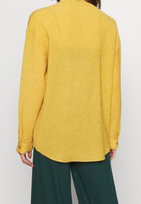 Camicia gialla con texture, colletto a bottoni, maniche lunghe e orlo curvo, abbinata a pantaloni verdi a gamba larga.