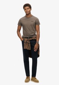 Brun kortærmet t-shirt, mørk denim, ternet skjorte bundet om livet, tan sko, sølvur. Glat stof, afslappet pasform, moderne design.