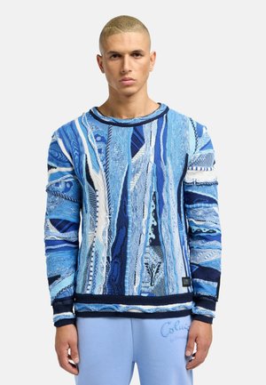 Blå mønsteret sweater med forskellige teksturer og nuancer, med rund halsudskæring og ribbede manchetter, parret med lyseblå bukser.