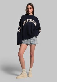 Maglione a coste blu navy con testo "WRSTBHVR", abbinato a una gonna in jeans sfrangiata e sneakers beige con plateau. Occhiali trasparenti completano il look.