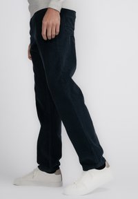 Donkerblauwe corduroy broek met een slim fit, voorzien van een zijzak en gedragen met witte sneakers. Het materiaal heeft een gestructureerd ribbelpatroon.