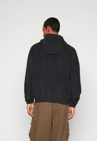 Moški stoji obrnjen stran, oblečen v črno kapuco iz flisa in ohlapne rjave cargo hlače proti enotni podlagi.