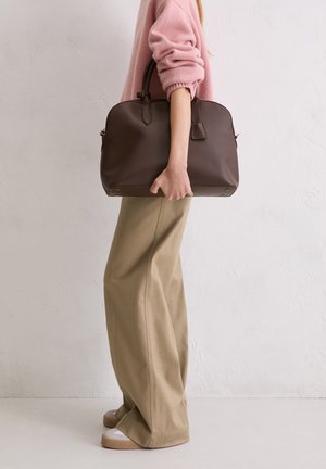 Person in beige wide-leg pants and pink sweater holding large brown leather handbag by its handles at side.
