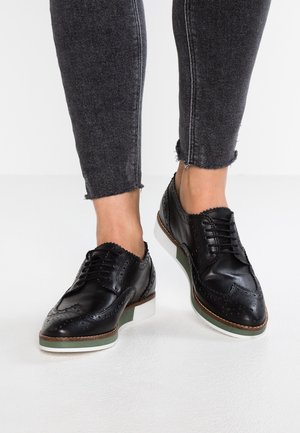 Schwarze Leder-Herrenschuhe mit Brogue-Details, Schnürverschluss vorne und einer kontrastierenden grünen und weißen Sohle. Kombiniert mit grauen Skinny-Jeans.