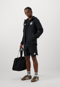 Musta urheiluasu, johon kuuluu vetoketjullinen takki logolla, yhdistelevät shortsit ja ruskeat kengät, pitäen mustaa urhelukassia. Raidalliset sukat näkyvissä.