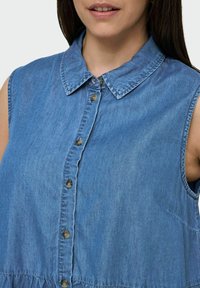 Denim bešėmis rankovėmis marškiniai su klasikiniu apykakle, priekiniais mygtukais ir surinkta juosmens konstrukcija. Medžiaga turi šviesiai mėlyną, tekstūruotą paviršių.