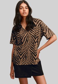 Femeie îmbrăcată cu un top scurt cu decolteu în V, cu dungi zebra maro și negre, și o fustă neagră, pozând cu o mână pe șold.