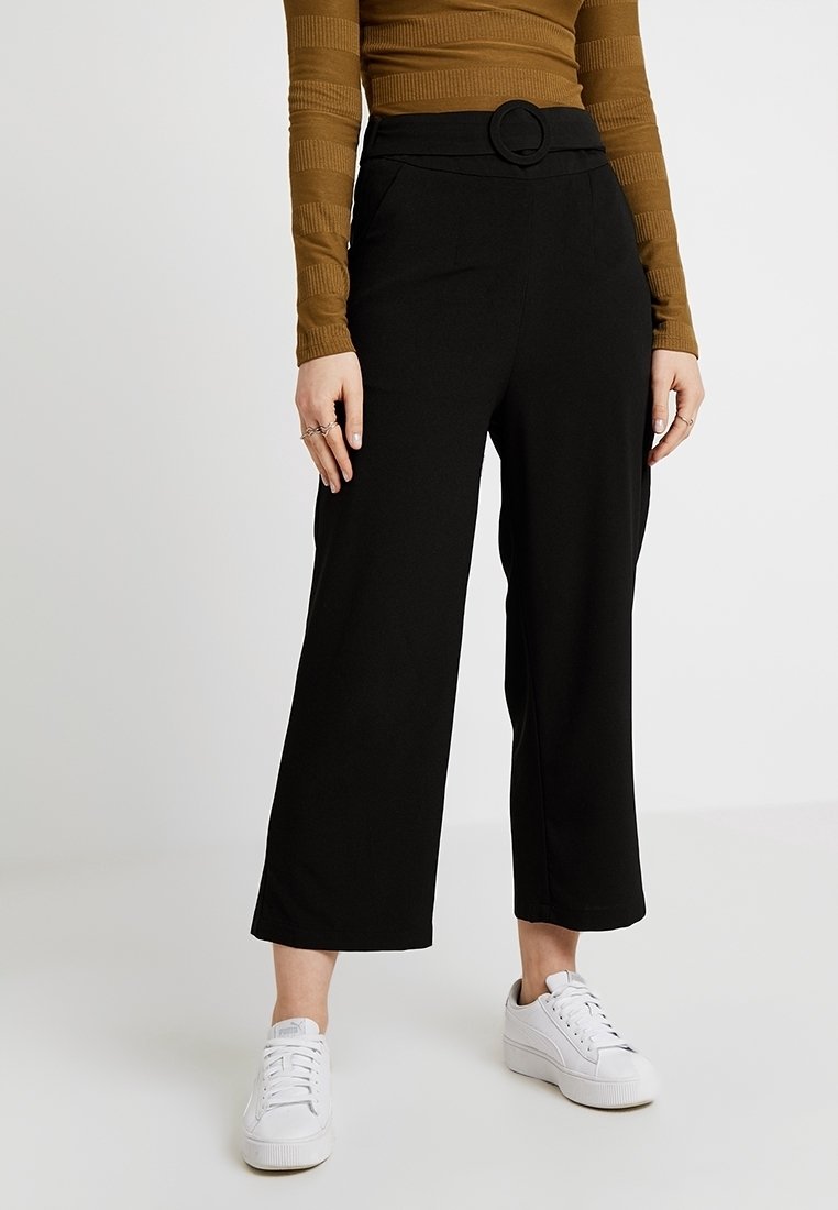 Svarta byxor med vida ben och hög midja, detaljerad med ett cirkulärt bälte, tillsammans med en ribbad brun skjorta och vita sneakers. Slät tygtexur.