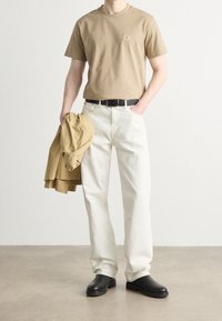 Ljusbrun bomullströja med en liten logotyp, vita denimbyxor, svart bälte, texturerade svarta stövlar och en beige jacka som hålls i handen.