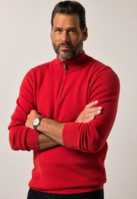 Roter Reißverschluss-Pullover mit hohem Kragen und langen Ärmeln, ausgestattet mit gerippten Bündchen und einer glatten Textur. Getragen mit einer braunen Armbanduhr.