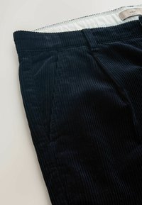 Un pantalon en velours côtelé navy présente une texture côtelée, une taille élastique, des poches latérales et une étiquette marquée "NEXT". Le tissu est doux et durable.