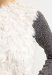 Faux fur-väst i vitt med en texturerad, fluffig yta, kombinerad med en grå långärmad topp i ett smidigt, stickat tyg.