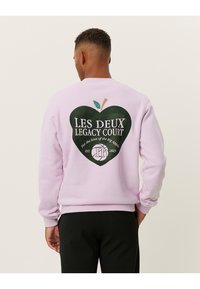 Λιλά φούτερ με σχέδιο σε σχήμα καρδιάς στο πίσω μέρος, με πράσινο και άσπρο κείμενο, και μια μικρή πράσινη λεπτομέρεια φύλλου από πάνω.