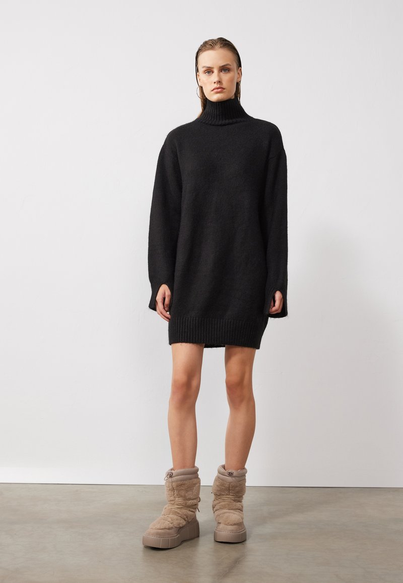 Robe pull en maille noire oversize avec col montant, présentant des finitions côtelées. Associée à des bottes de cheville beiges et rembourrées avec des semelles compensées.
