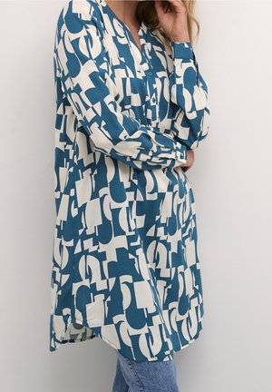 Frau trägt eine langärmelige, locker sitzende Tunika mit einem blau-weißen abstrakten geometrischen Muster, kombiniert mit hellblauen Jeans.