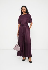 Macacão de cetim roxo com mangas curtas, cinto na cintura e pernas largas. Combinado com uma mala em tom cinza claro e sapatos de salto alto pretos.