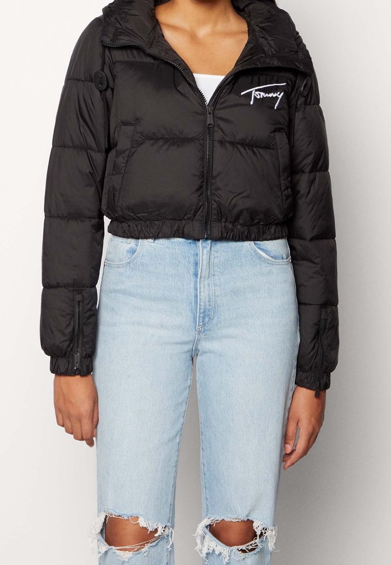 Černá péřová bunda s cropped designem, lesklým materiálem, zipem vpředu, kapucí a elastickými manžetami. Na hrudi má logo Tommy.