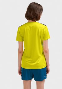 Kortärmad, ljusgul idrottströja med svarta detaljer vid axlarna, tillsammans med blå shorts. Slät struktur och avslappnad passform.