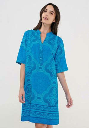 Mujer con un vestido azul suelto hasta la rodilla con patrones geométricos y florales, de pie contra un fondo claro y liso.