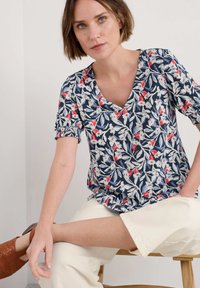 Ziedu raksta blūze tumši zila ar baltām un sarkanām akcentiem. V-veida kakla dizains, īsas pūkainas piedurknes ar savilktām aprocēm, maiga auduma tekstūra.