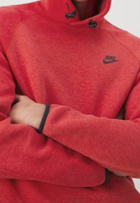 Person klädd i en röd Nike-hoodie med korslagda armar, som visar den svarta Nike-logotypen och detaljer i svart på ärmarna.